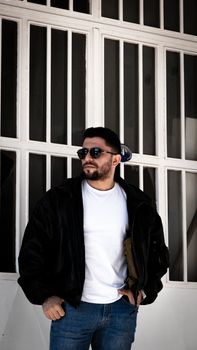 Stylish man with sunglasses wearing jacket and jeans against a white wall, exuding cool fashion vibe.