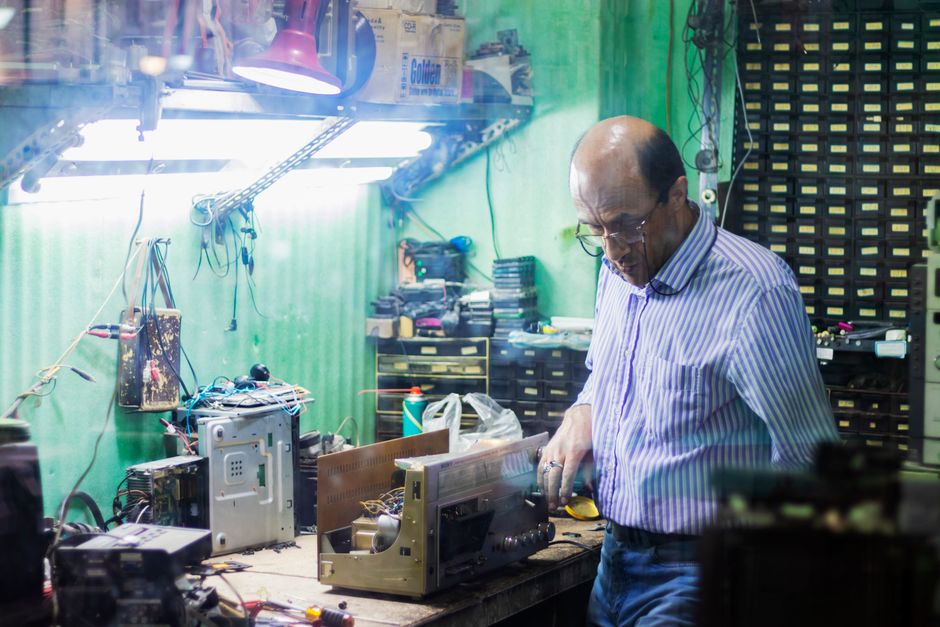 Man focusing on repairing electronic equipment in cluttered workshop with various tools.