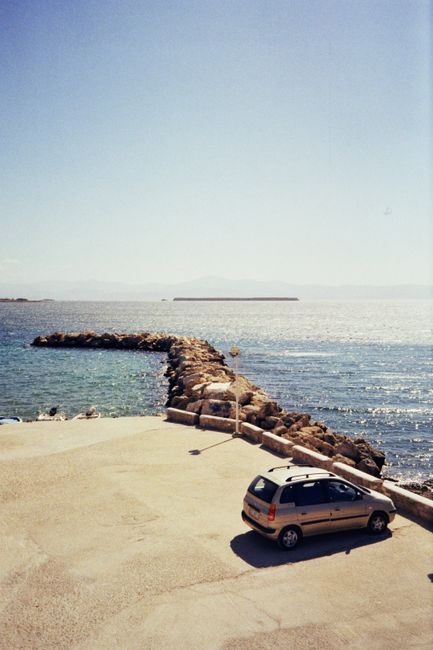 Captivating aerial view of Paros, Greece, featuring a car parked by the coastline and a stone breakwater extending into the sea.
