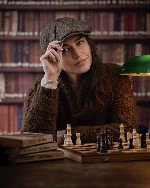 Smiling Woman in a Newsboy Cap and a Brown Cardigan Sitting at a Chessboard in the Library