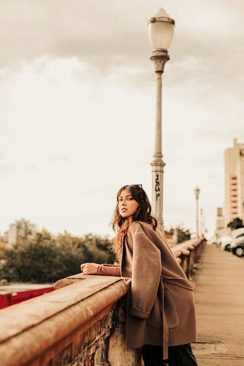 Young Woman in a Coat Standing on a Bridge in City 