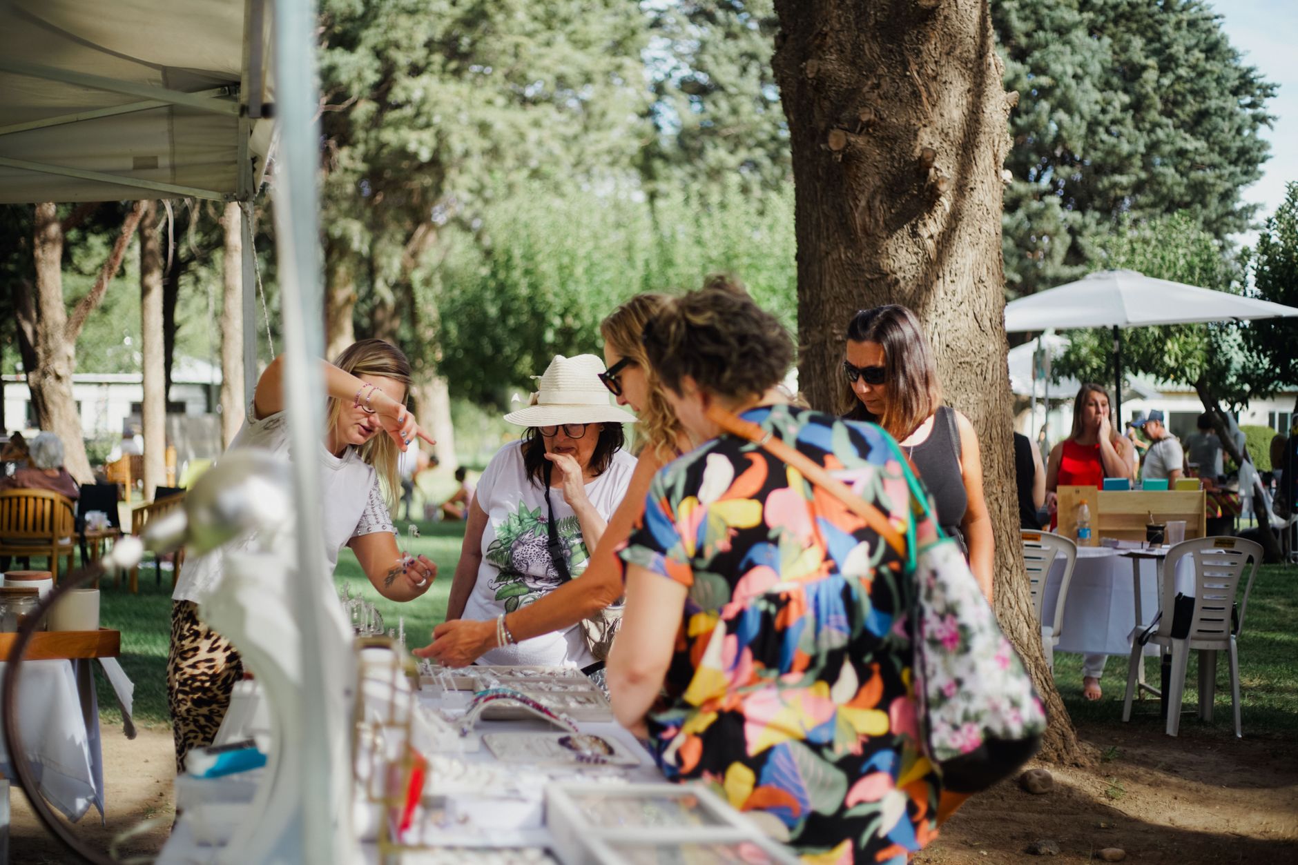 A vibrant outdoor market scene featuring shoppers exploring a jewelry stand under the shade of trees.