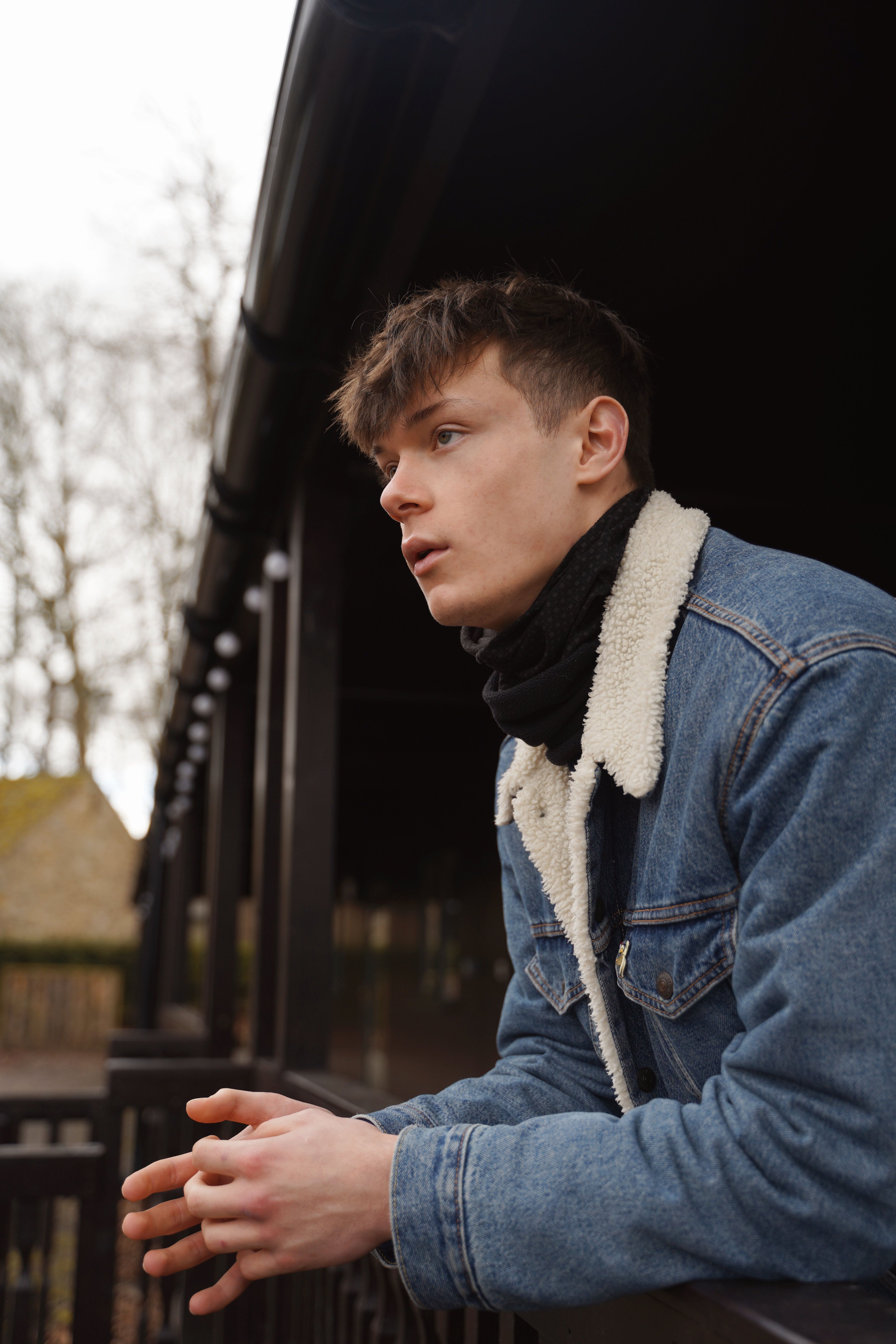 A young man in a denim jacket leans on a balcony railing, gazing thoughtfully.
