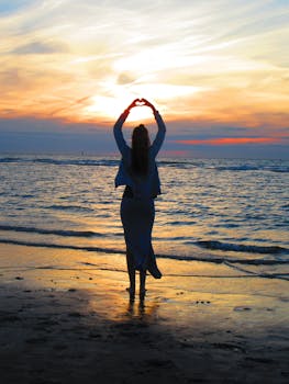 Woman With Arms Up Making Heart Sign While Standing on Beach at Sunset