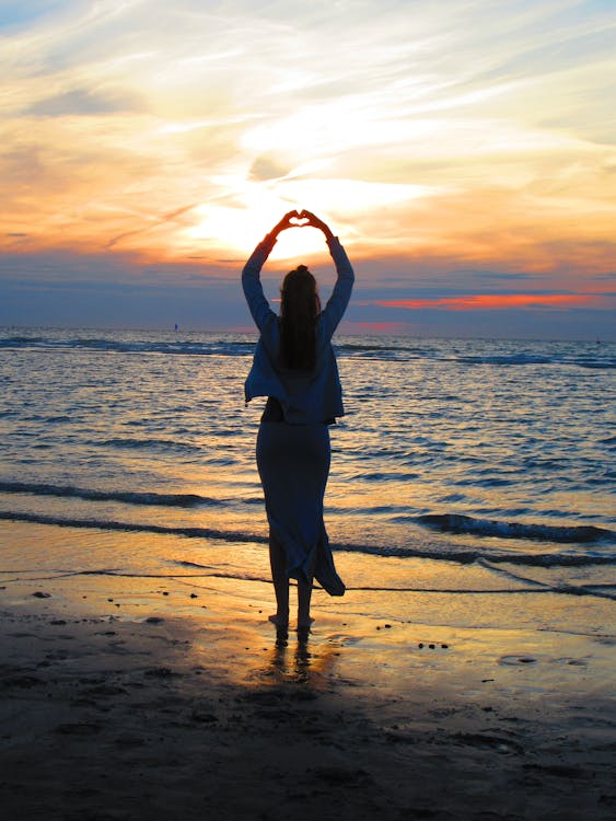 女人，雙臂高舉使心臟標誌，而站在海灘上在日落時