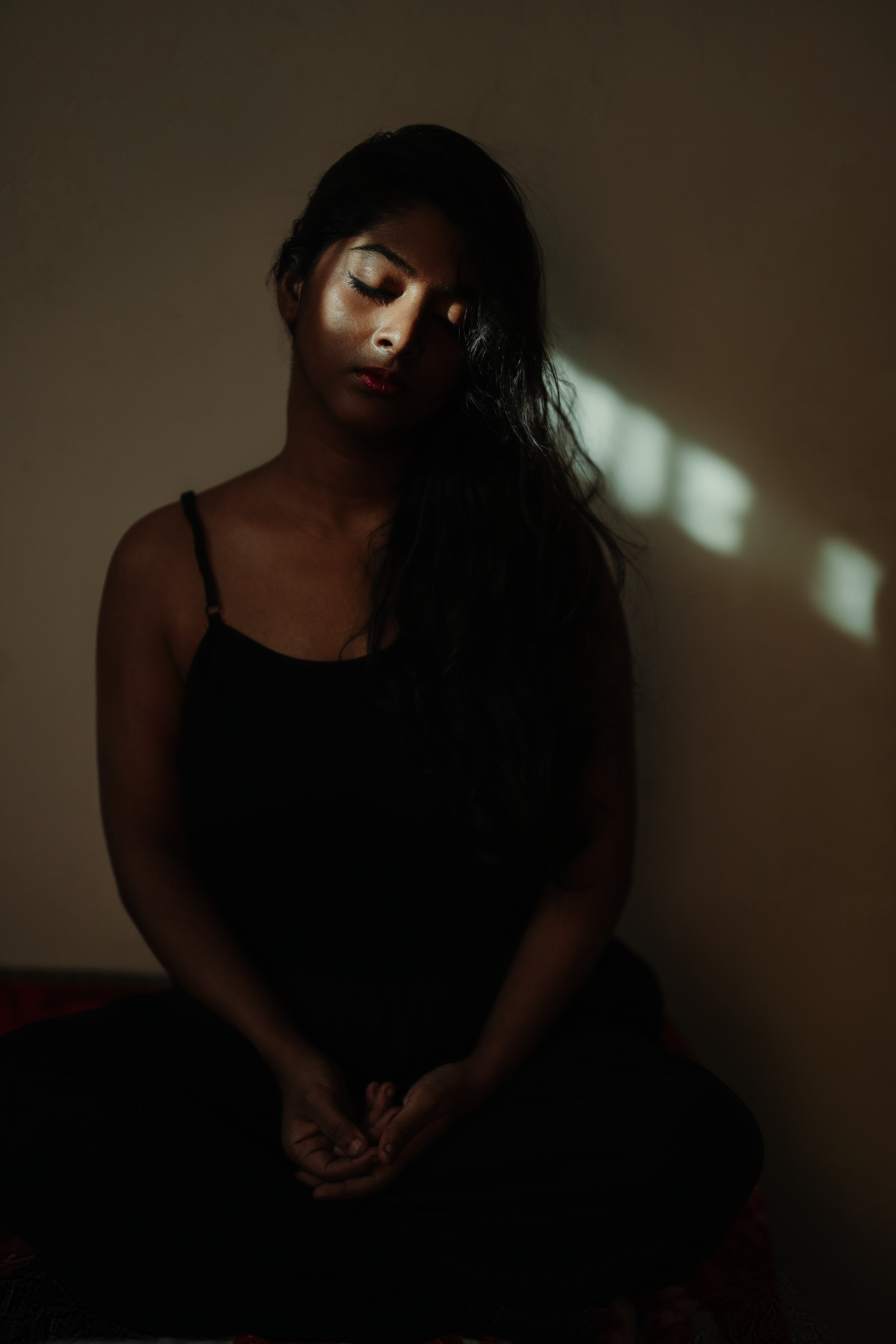 A woman meditates in a dark room with soft sunlight casting shadows.