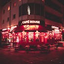 一个热闹的咖啡馆，点缀着胭脂色与红色霓虹灯，并设有户外座位，夜景完美地营造出充满活力的夜生活氛围