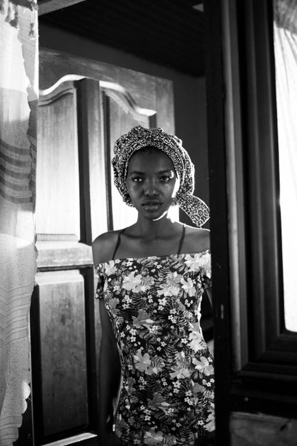 Black and white portrait of a woman in a floral dress and headscarf standing by a doorway.