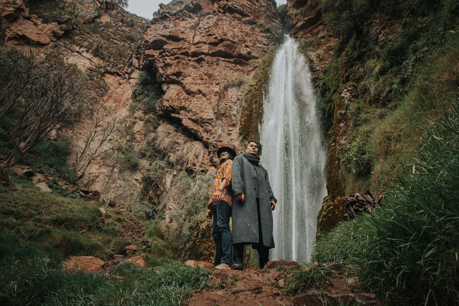 Two fashionable men posing near a majestic waterfall in Ollantaytambo, Peru.