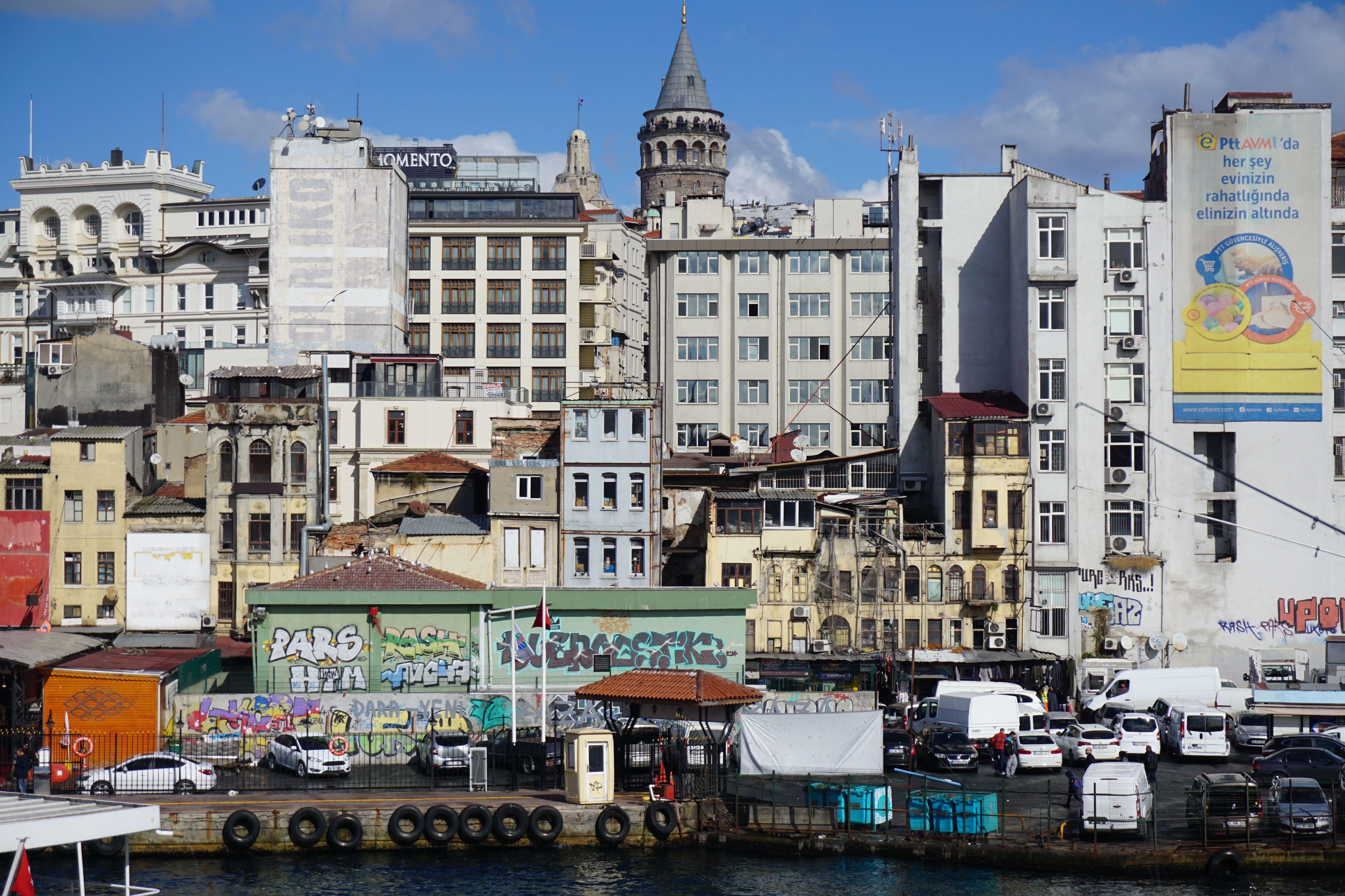A waterfront with moored boats and a graffiti-covered wall below crowded apartment buildings, with a stone tower with a conical roof rising behind