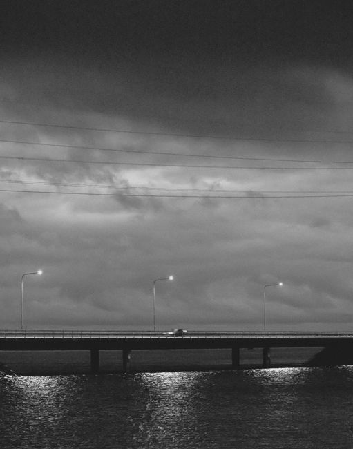 A moody black and white photo of a bridge over water under cloudy night skies.