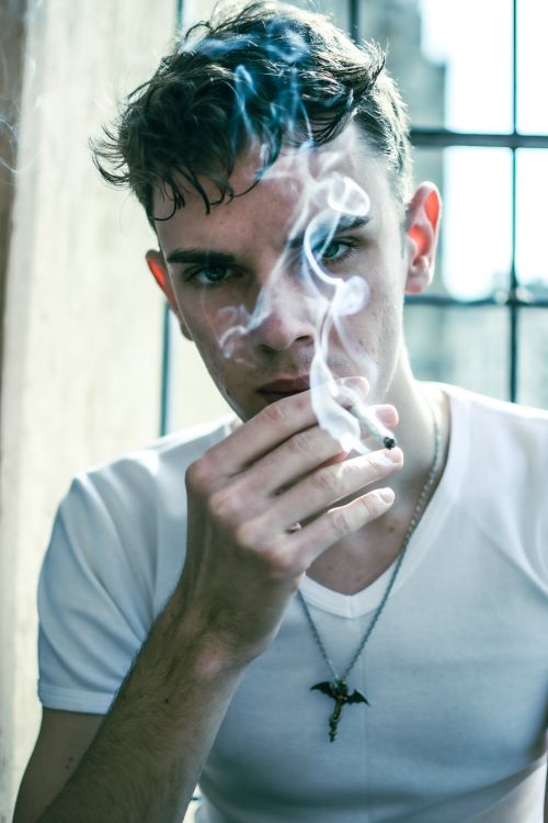 Free Close-up portrait of a young man smoking, showcasing modern fashion style with a necklace. Stock Photo