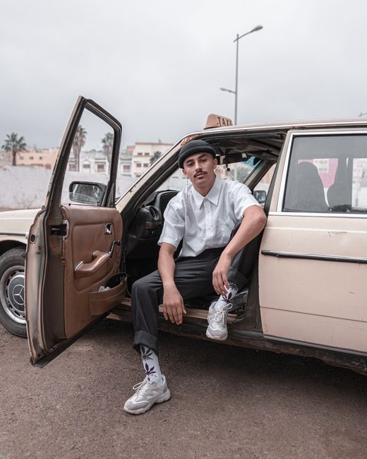 A stylish young man wearing casual attire sits in an old car parked outdoors in Morocco.
