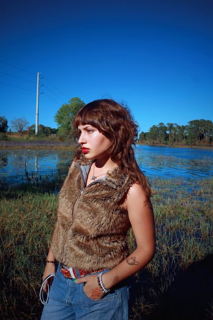 Stylish young woman posing by a scenic lake, showcasing autumn fashion with fur vest and denim.