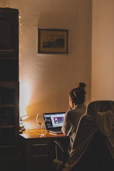 Woman working on a laptop at a dimly lit desk in a cozy room, viewed from behind.