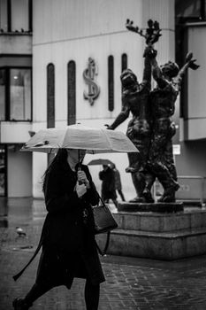 Black and white photo of a woman with an umbrella walking in Ankara near a sculpture.