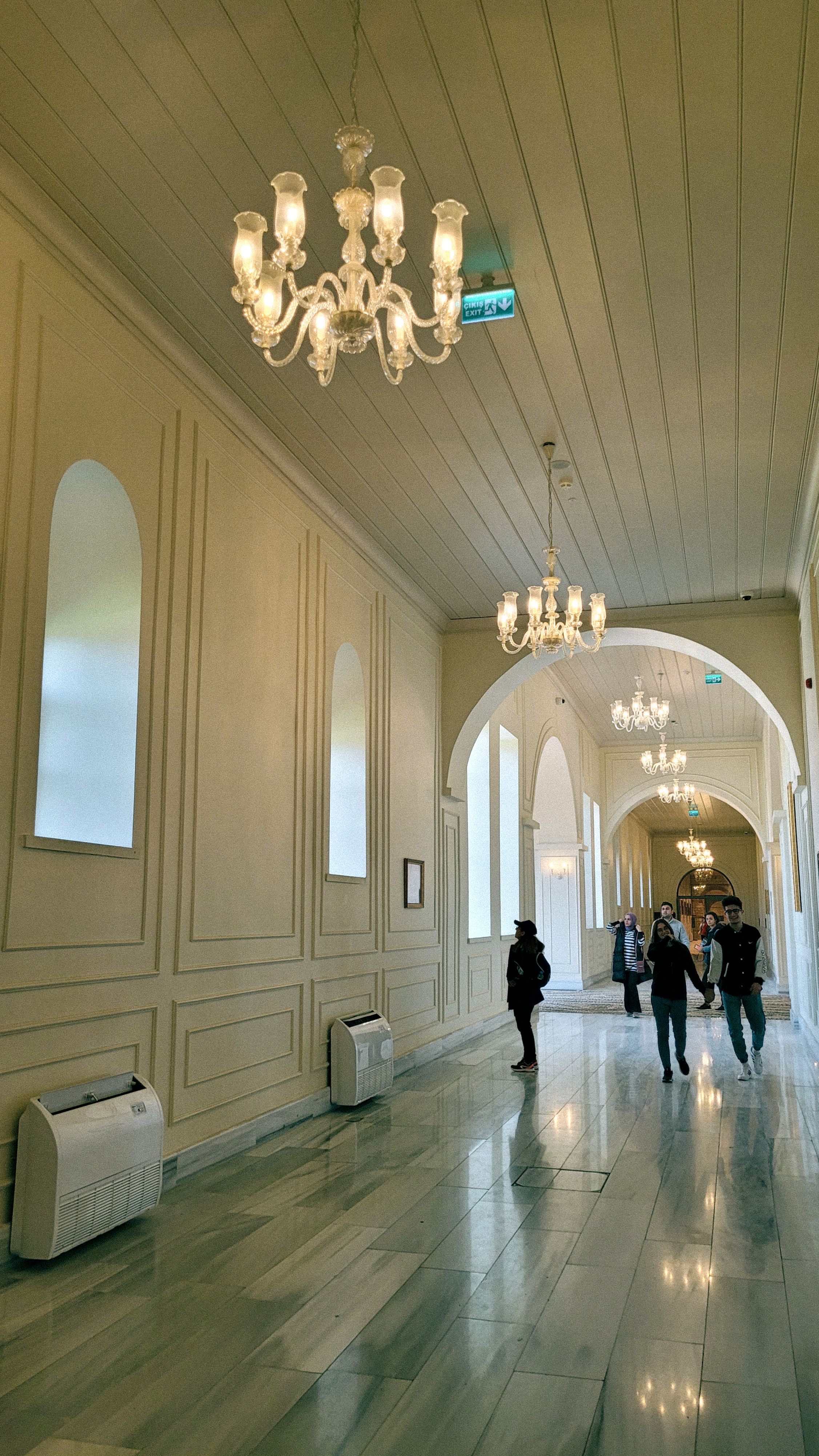 A luxurious museum corridor featuring elegant chandeliers and visitors exploring the space.