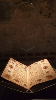 An ancient illuminated manuscript opened and displayed under focused lighting in dim surroundings.. ancient, illuminated, man