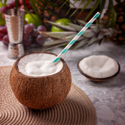 Free Close-up of a coconut drink with a striped straw on a table, surrounded by tropical fruits. Stock Photo