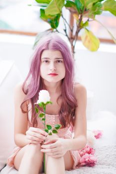 A young woman with pink hair holds a flower indoors, exuding a soft, elegant charm.