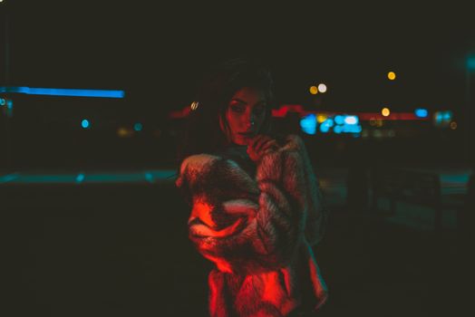 Stylish woman in fur coat under moody red lights at night.