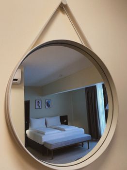 A round mirror reflects a modern, minimalist bedroom with a bed and art on the wall, creating a serene feel.