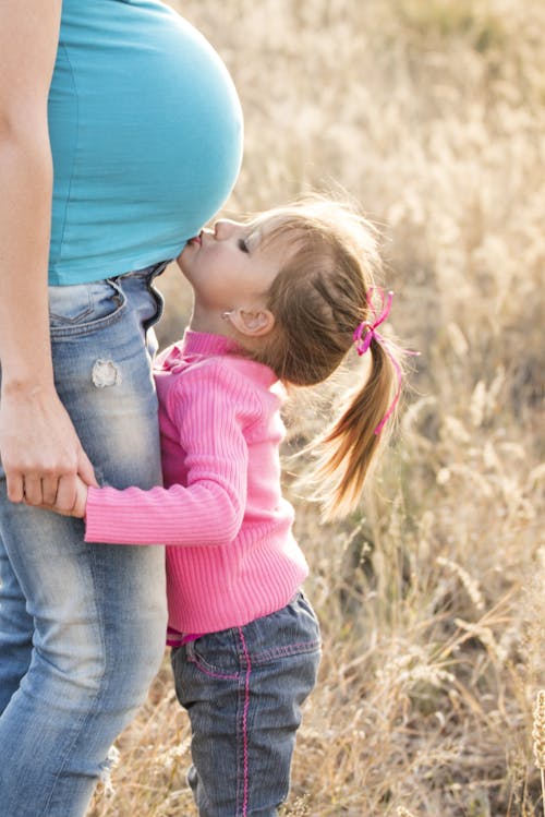 Gadis Dengan Sweater Merah Muda Dan Jeans Abu Abu Berciuman Perut Wanita Hamil Dengan Kemeja Biru Dan Jeans Denim Biru