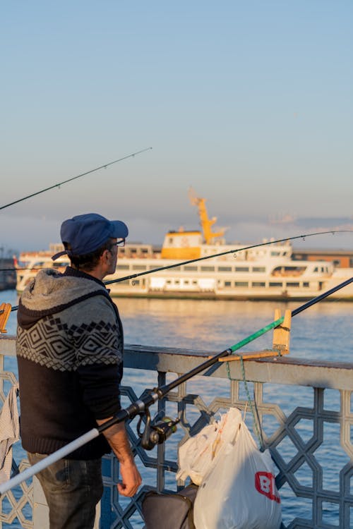 A man is fishing on a pier with a boat in the background