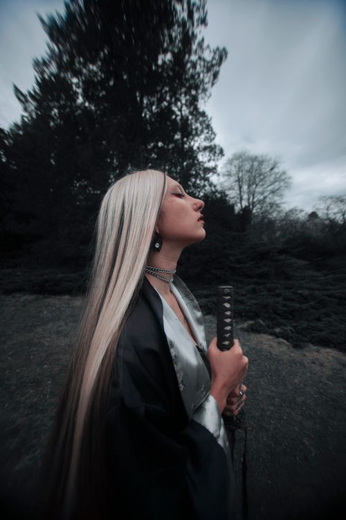 Free A woman with long blonde hair and a sword poses elegantly outdoors, surrounded by trees. Stock Photo