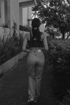 Monochrome photo capturing a woman walking outdoors on a pathway lined with plants.