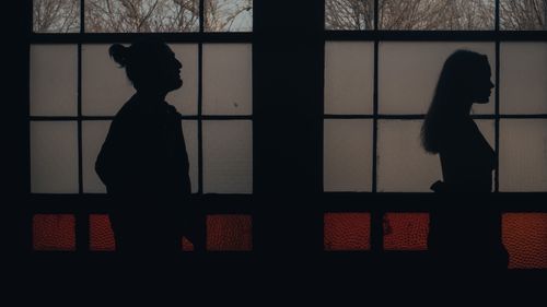 Free Silhouettes of a man and a woman standing apart indoors, backlit by a window. Stock Photo