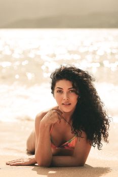 A woman with curly hair in a colorful swimsuit posing on a sunny beach shore.