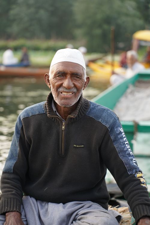 Free A senior adult man in a knitted sweater and cap sits smiling on a boat by the water. Stock Photo