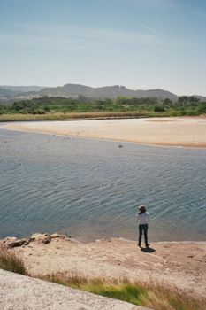 A woman stands by a peaceful riverbank, capturing the serene landscape of nature.