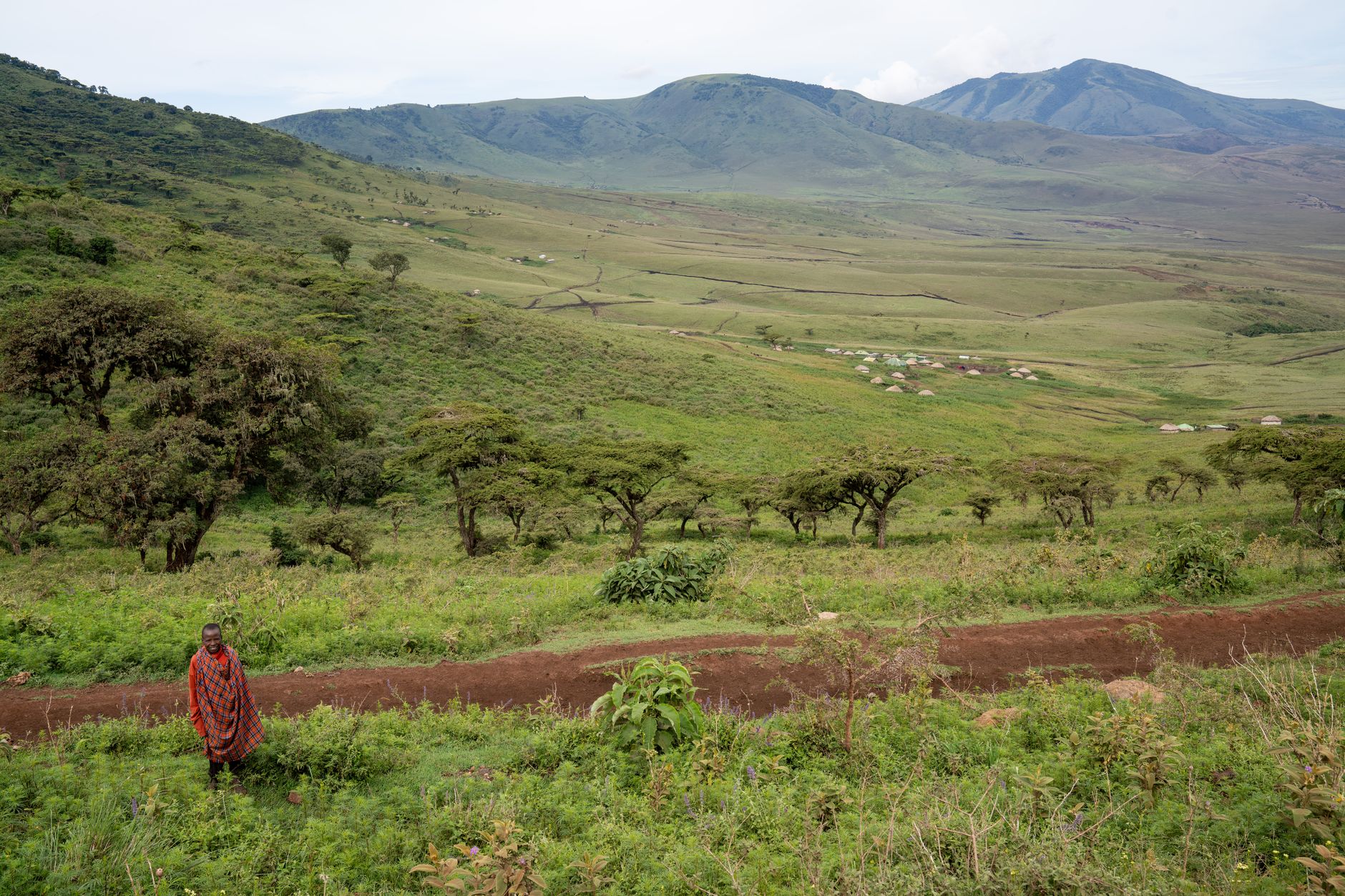 A Maasai individual stands by a dirt road amidst lush green hills and scenic landscape.