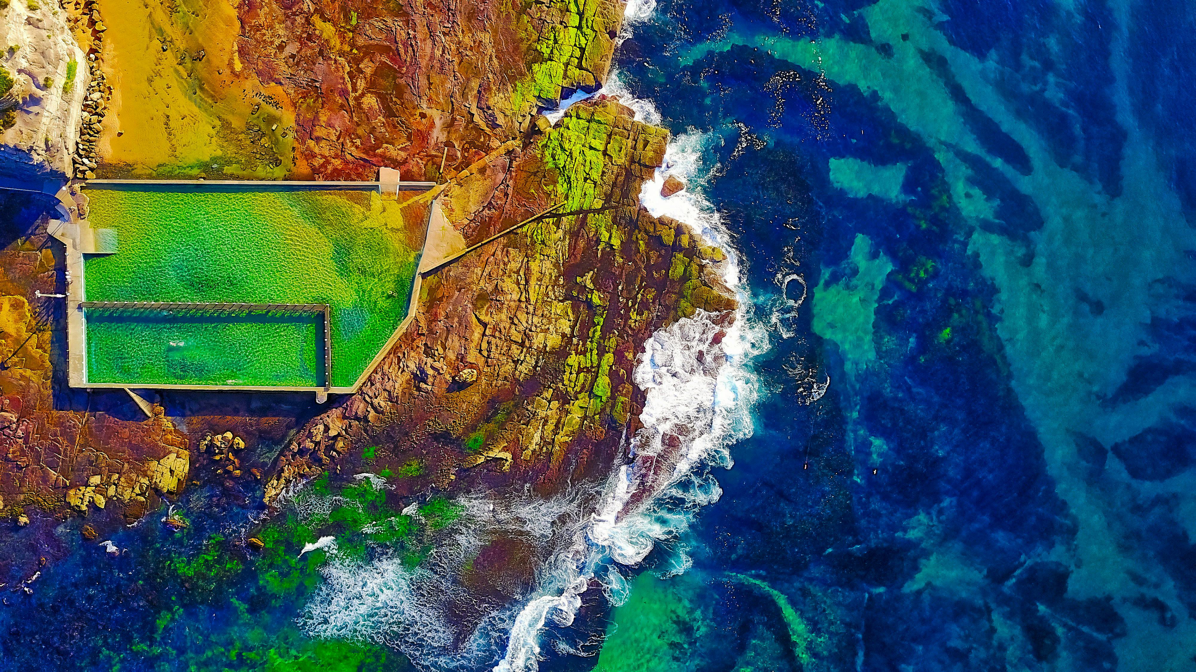 一张令人叹为观止的鸟瞰图，展现了一个充满活力的沿海游泳池，正对着五颜六色的岩层和广阔的公海