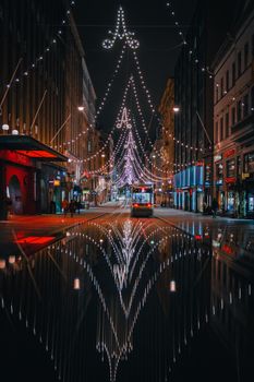 Captivating night scene of a Helsinki street adorned with Christmas lights and a tram reflecting on wet pavement.