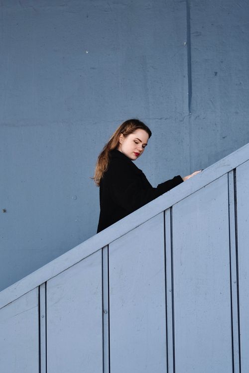 Free Young woman in black coat walking up stairs with blue background. Stock Photo