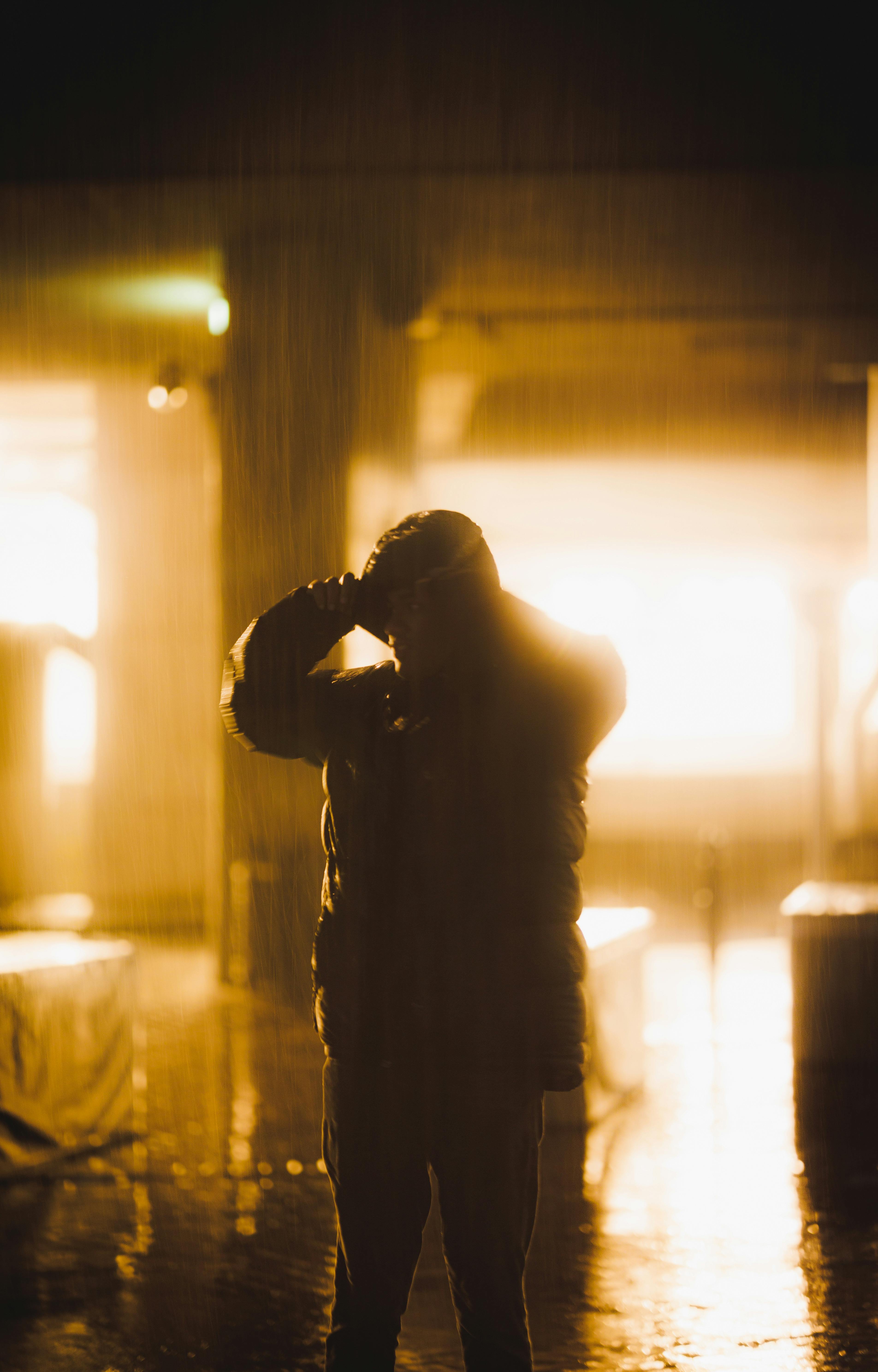 A silhouette of a person wearing a jacket standing in a backlit rainy city scene at night.