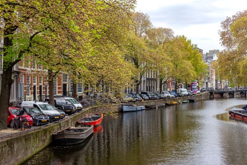 Free Serene view of Amsterdam canal with boats, cars, and lush autumn trees. Stock Photo