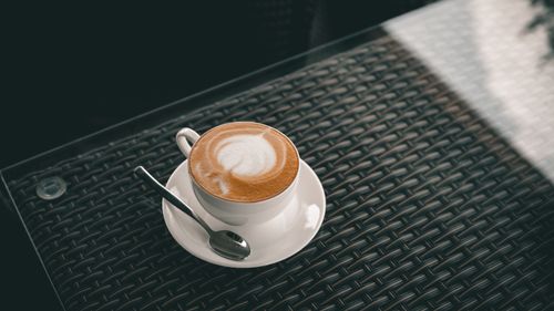 Free A high-angle shot of a cup of latte with intricate froth art served on a woven rattan table. Stock Photo
