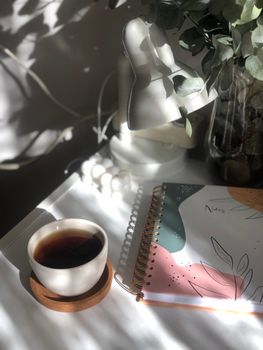A cozy desk setup with coffee, notebook, and greenery, captured in soft lighting.