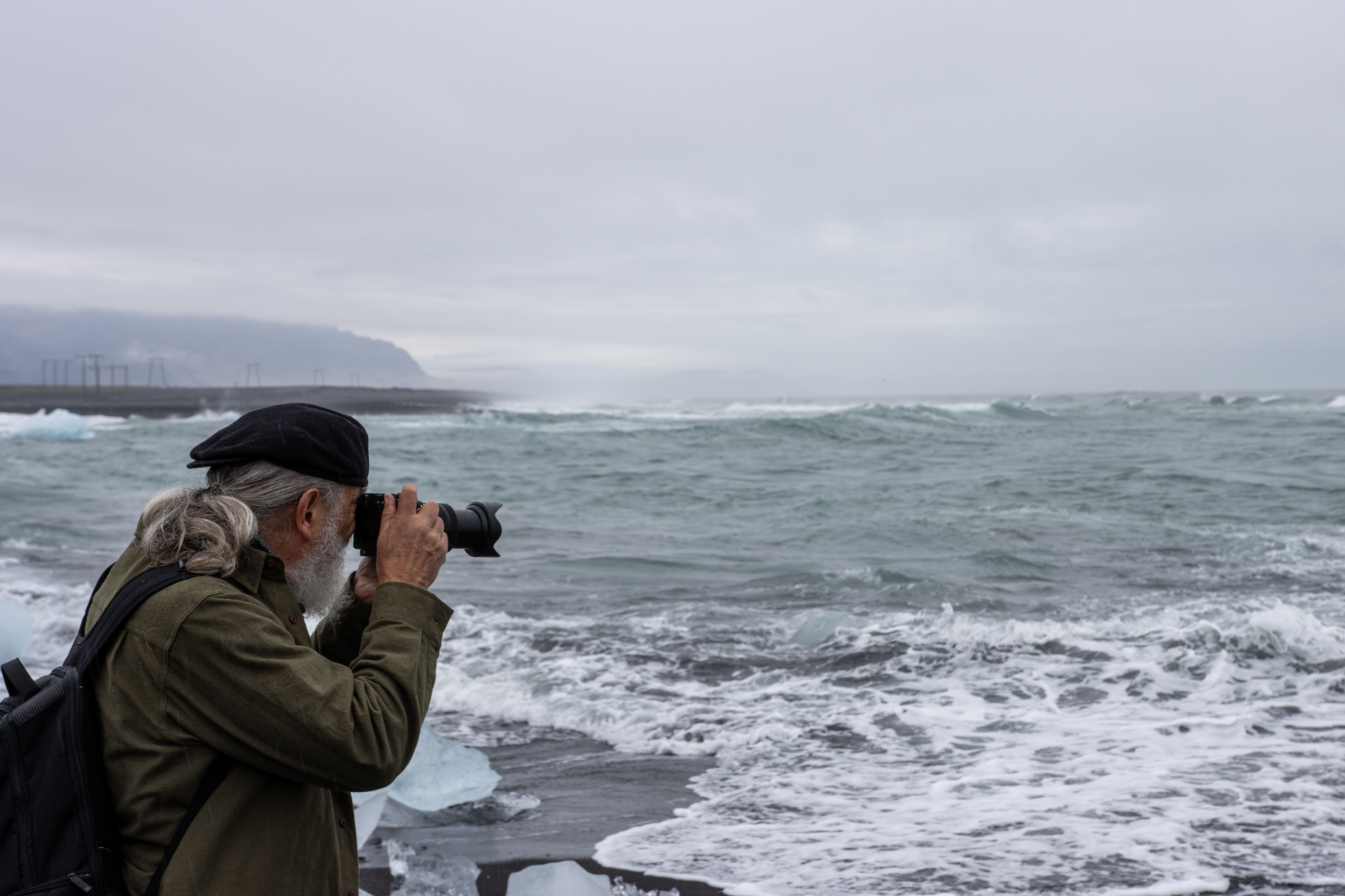 Elderly man photographs stormy coastline at a seashore, capturing dramatic waves.