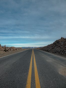 Long open road stretching to the horizon beneath a cloudy sky, surrounded by rocky terrain.