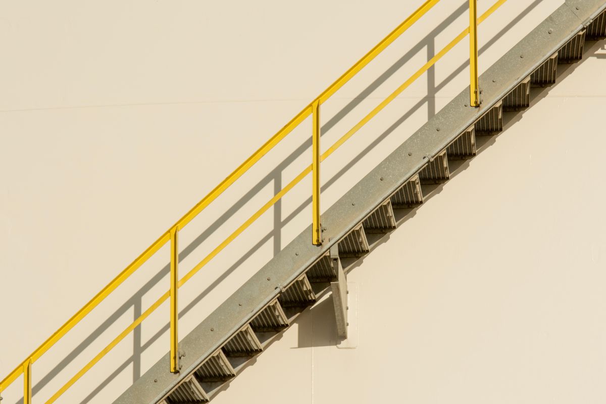 Scară metalică industrială pe diagonală, cu balustradă vopsită în galben aprins, pe un perete neutru