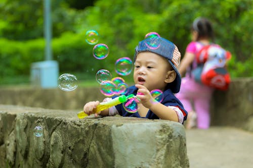 бесплатная Мальчик в синей кепке играет мыльные пузыри и прислоняется к серой бетонной стене днем Стоковое фото