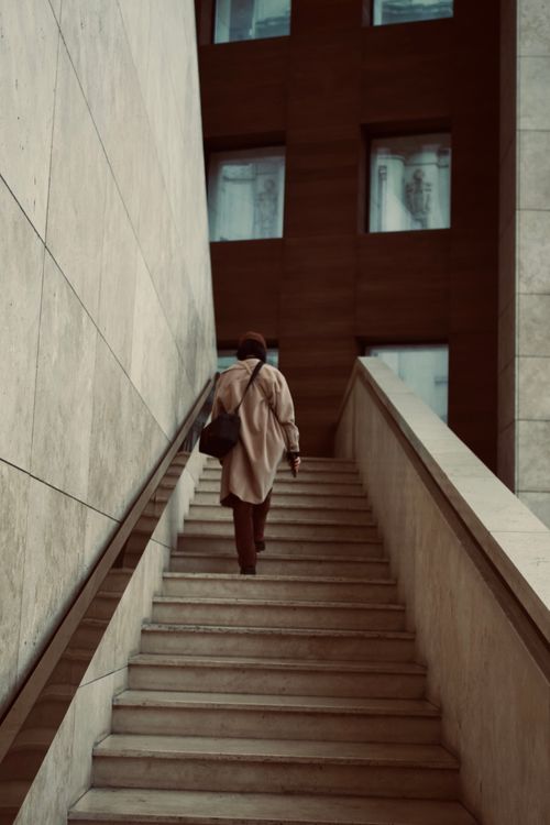 Free A woman in a coat walks up marble stairs, surrounded by urban architecture. Stock Photo