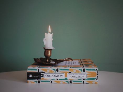 A lit wax candle on a brass holder rests atop the book Oblomov against a green backdrop.