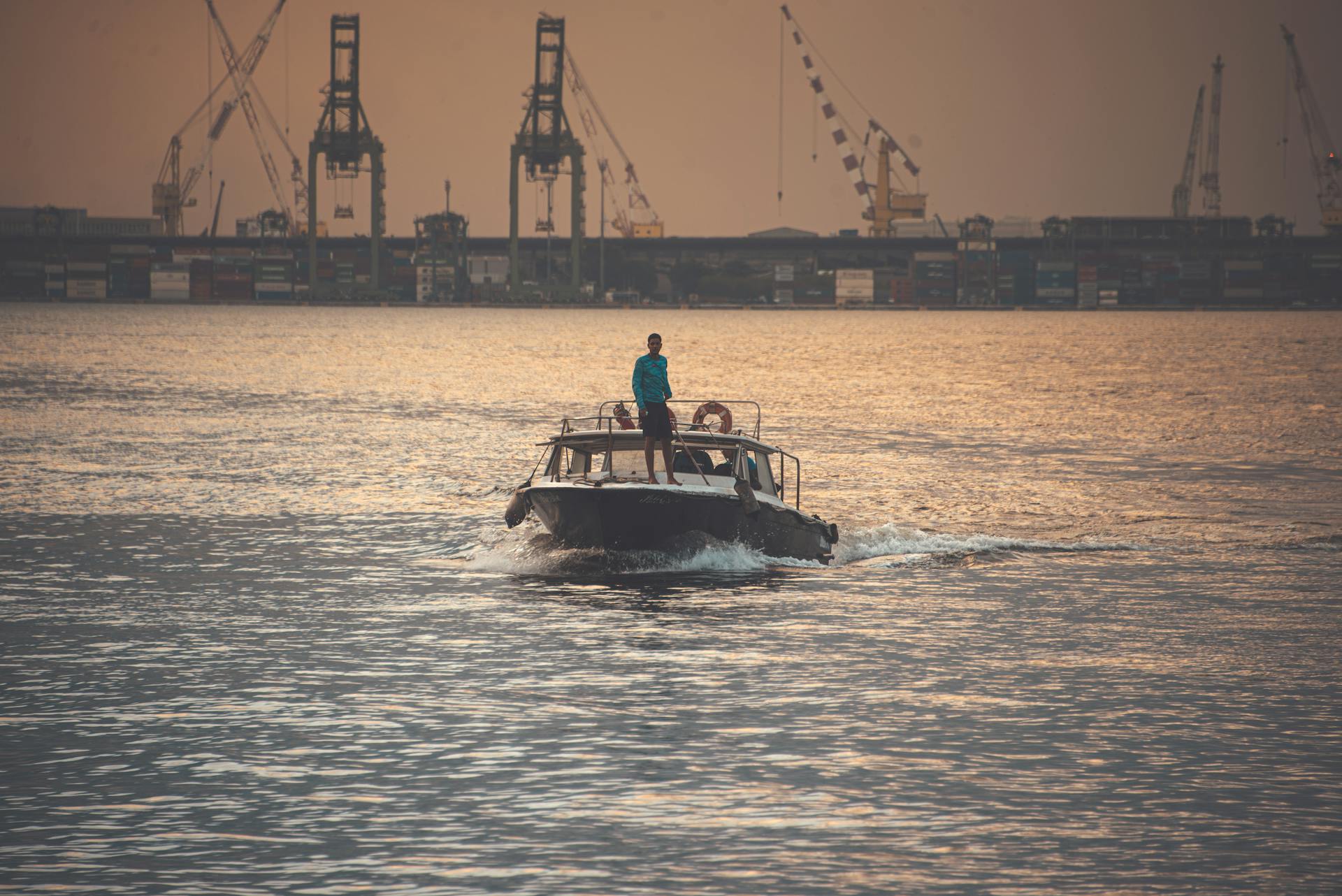 A motorboat navigating the waters near a port in Rio de Janeiro at sunset, with industrial cranes in the background.