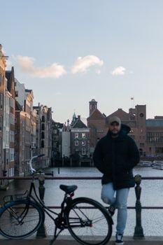 A man standing by a canal with a bicycle on an Amsterdam bridge, showcasing urban architecture.